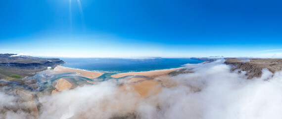 Aerial view of Rauðasandur, Iceland, where golden sands meet turquoise waters, framed by dramatic cliffs and low-hanging clouds under a radiant blue sky.