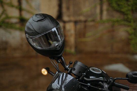 A sleek black motorcycle helmet with rain drops, emphasizing protection and style, sits on a bike in an urban setting under a rustic backdrop.