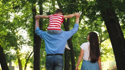 Father rocks child sitting on shoulders among trees in city park. Grateful mother and father stroll...