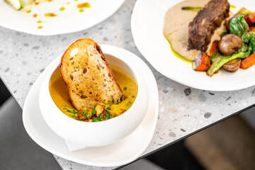 A bowl of creamy soup garnished with fresh herbs and a toasted bread slice, served on a white plate. In the background, a meat dish with vegetables is visible on a speckled countertop.