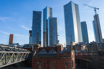 Obraz premium View of Manchester, England, showing Deansgate Square skyscrapers, a red brick Victorian building, a pedestrian bridge, and a construction crane.