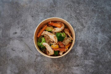 A bowl of food with vegetables and chicken. The bowl is made of paper