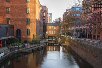 Naklejka premium A canal in Manchester, England, with a central lock, historic red brick buildings, modern structures, colorful flags, and outdoor seating areas.