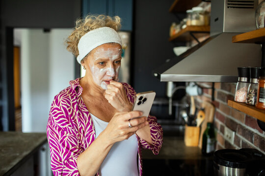 Worried senior woman with face mask looking at smartphone in kitchen