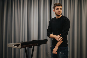 Young man in casual clothes poses next to an electronic keyboard in a studio environment, evoking...