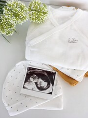 A baby ultrasound photo next to soft white baby clothes and hats, symbolizing pregnancy and preparation for a newborn.