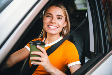 Smiling young woman in casual orange t-shirt holding coffee cup inside a car on a sunny day...