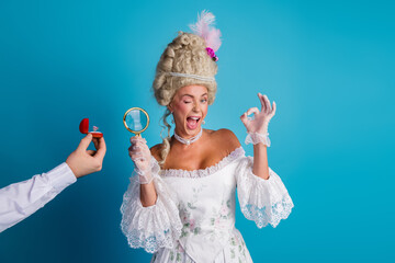 Fototapeta premium Elegant woman in baroque attire humorously examining a ring, blending historic and modern elements in a vibrant setting