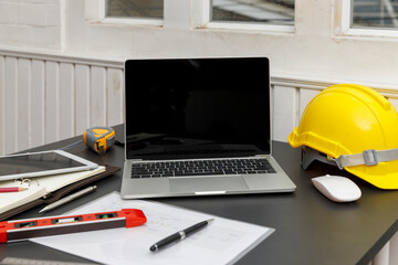 A laptop with a blank screen sits beside a yellow hard hat on a desk, suggesting a blend of technology and construction or engineering.