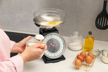 Woman holding spoon with flour near mechanical kitchen scale at white marble countertop indoors,...