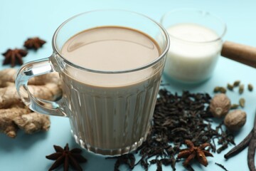 Aromatic Masala tea in glass cup, spices, dry leaves and milk on light blue background, closeup