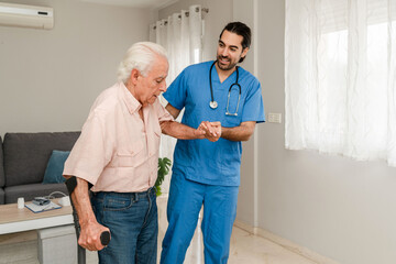 Fototapeta premium Kind male nurse in blue uniform helping a senior man with crutches during home visit
