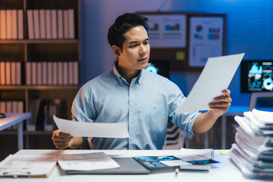 Asian businessman analyzing paperwork and comparing financial reports while working late in office at night