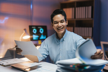Fototapeta premium Young office worker comparing documents and smiling while working late in the office with computer and paperwork