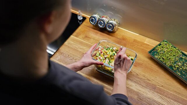 Over shoulder shot: professional chef carefully mixing vibrant seasonal salad in transparent glass bowl, arranging fresh ingredients on rustic wooden surface with herb jars and green sprouts nearby