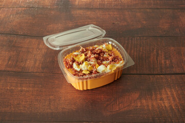 A container of food with a lid on top. The lid is plastic. The food is yellow and has bacon and eggs in it