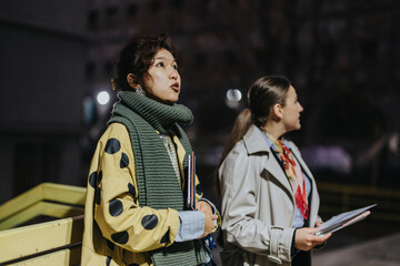 Two women engage in discussion outdoors during nighttime. The scene portrays communication, urban life, and connection between people in a casual setting.