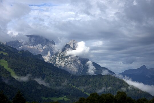 The image captures a breathtaking view of a mountain range shrouded in mist. Lush green forests cover the slopes, with wispy white clouds swirling and drifting through the valleys. The scene is bathed