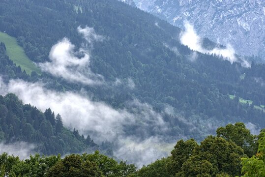 The image captures a breathtaking view of a mountain range shrouded in mist. Lush green forests cover the slopes, with wispy white clouds swirling and drifting through the valleys. The scene is bathed