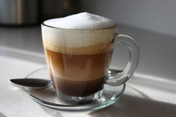 A beautifully layered coffee in a clear glass, displaying varying shades of brown topped with frothy cream, sitting on a countertop