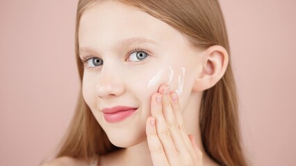 Portrait of a smiling cheerful girl applying face cream on cheek for skin glow. Advertising face cream and skincare products for teens. Blonde girl with blue eyes isolated in studio on pink background