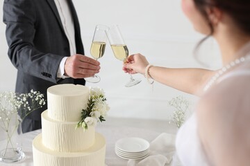 Married couple clinking glasses of champagne at table with wedding cake indoors, closeup