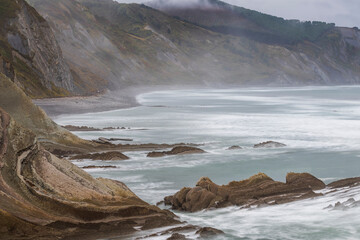 Powerful waves crashing against rugged flysch cliffs at Itzurun Beach, Cantabria, generating dramatic moody seascape under overcast northern Spanish skies