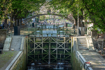 Paris, France - 04 05 2025: Canal Saint-Martin. Panoramic view of people sited, apartment buildings, a bridge and trees reflecting on the water.