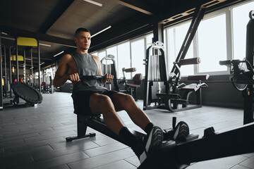 Handsome male practicing on rowing machine in fitness gym. Athletic young man working out on rowing machine at fitness center. Weight reduction and cardiovascular workout concept. © Davidovici
