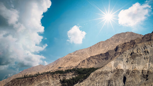 An alpine landscape of Himalayan rocky mountain ranges, with forest trees under bright sunlight and a cloudy blue sky in Gilgit-Baltistan, Pakistan. - Powered by Adobe