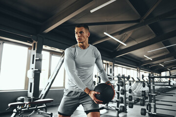 Horizontal photo of athletic man model exercising with medicine ball at gym. Young strong guy doing workout using fitness ball. Strength training, discipline, and fitness lifestyle concept
