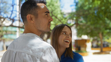 Fototapeta premium Couple enjoying a sunny day in an urban park with lush trees, highlighting a sense of love and togetherness between a smiling woman and man surrounded by nature.