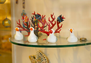 Four small white Murano glass bird figurines with orange beaks and red crests sit on a glass shelf with red Murano glass coral and colorful Murano glass fish behind them