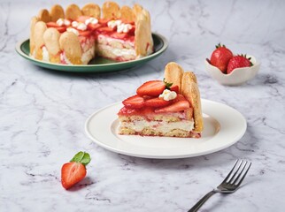 Sliced ​​round tiramisu cake with fresh strawberries and mascarpone cream decorated with marshmello flowers on a green plate on a light marble background. No-bake desserts. Italian cuisine