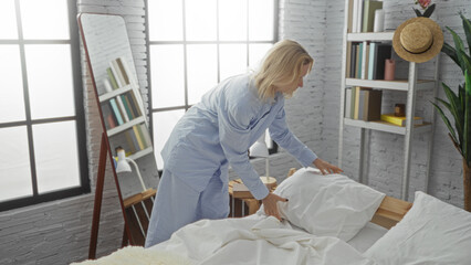 Woman making bed in cozy bedroom with mirror bookshelf and window light streaming in wearing blue...