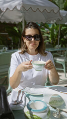 Woman enjoying coffee on a luxury restaurant terrace, wearing sunglasses and a white outfit, surrounded by stylish decor and natural greenery under sunlight.