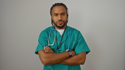 Young man with braids in green scrubs and stethoscope stands confidently against white wall, embodying professional afro-latino healthcare representation with arms crossed.