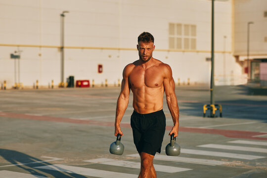 Muscular athlete walking and carrying two kettlebells in a parking lot