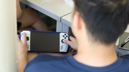 Close-up of a person playing on a handheld gaming console with blank screen, seated indoors near a table with another person in the background. © Timothy Figueroa