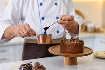 Elderly pastry chef with scoop and spoon of melted chocolate making cake at table in kitchen, closeup