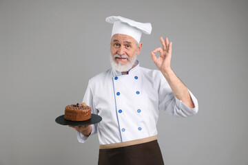 Professional pastry chef with delicious chocolate cake showing ok gesture on grey background