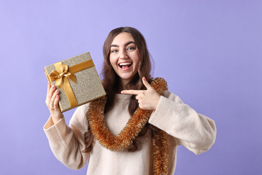 Happy young woman with tinsel and gift box on purple background