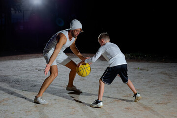 A father and son enjoy a night basketball game at a local park under bright lights focusing on teamwork and fun