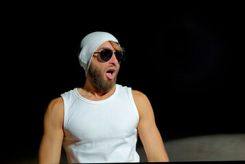A man wearing a tank top, sunglasses, and a beanie poses confidently at night against a dark...