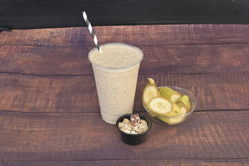 A cup of smoothie with a straw in it sits on a wooden table. A bowl of fruit is next to the cup