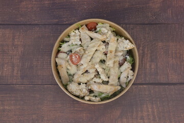 A bowl of salad with chicken and tomatoes. The bowl is on a wooden table. The salad is served in a plastic container