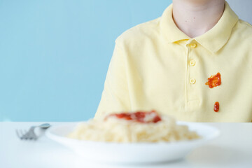Dirty drops tomato sauce or ketchup stain on yellow clothes during lunch. The setting features a simple blue wall, creating a casual dining atmosphere. Space for text