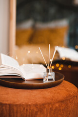 Liquid home fragrance in glass bottle with bamboo sticks with open paper book over glowing lights in bedroom closeup. Cozy home interior. Aromatherapy.