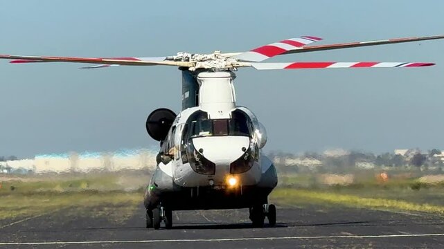 Head-on view of a large firefighting, twin-rotor helicopter taxiing and barely lifting off an airport runway while helmeted crewmembers lean out of its windows.