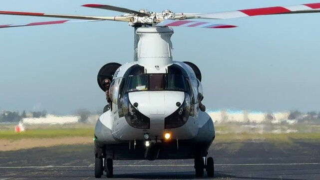 Head-on view of a large firefighting, twin-rotor helicopter taxiing forward on an airport runway while helmeted crewmembers lean out of its windows.